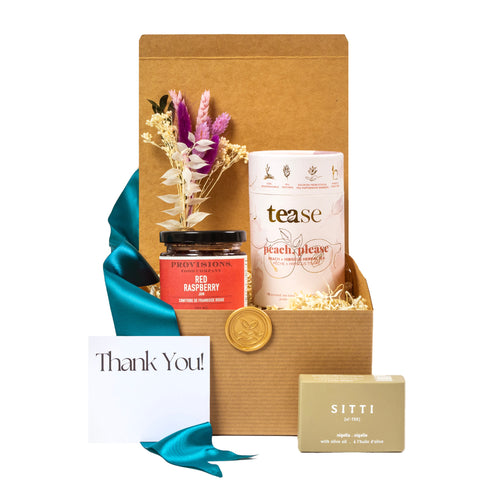 Thoughtful eco-friendly gift basket featuring red raspberry jam, peach herbal tea, olive oil soap, dried floral arrangement, and a thank-you card, all packaged in a sustainable cardboard box with teal ribbon. Ideal for corporate gifting or personal appreciation